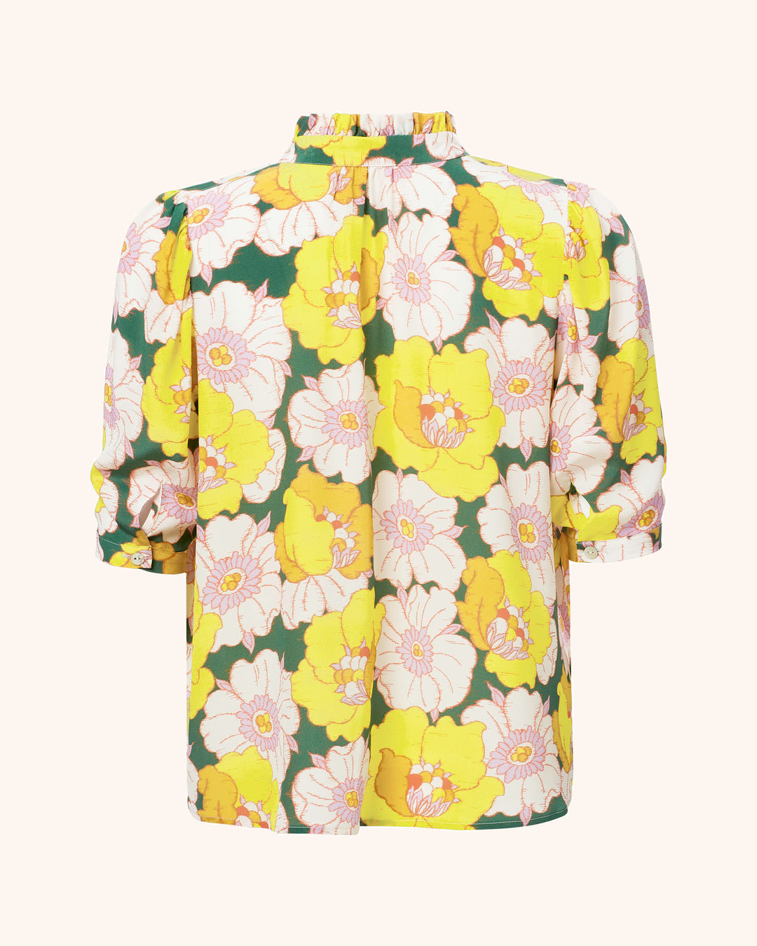 The Winnie Citrus Blossom Silk Shirt is a Liberty-print floral blouse made from Italian silk, featuring a ruffled high neck and quarter-length sleeves. The design displays large yellow and white flowers with green leaves set against a soft beige background.