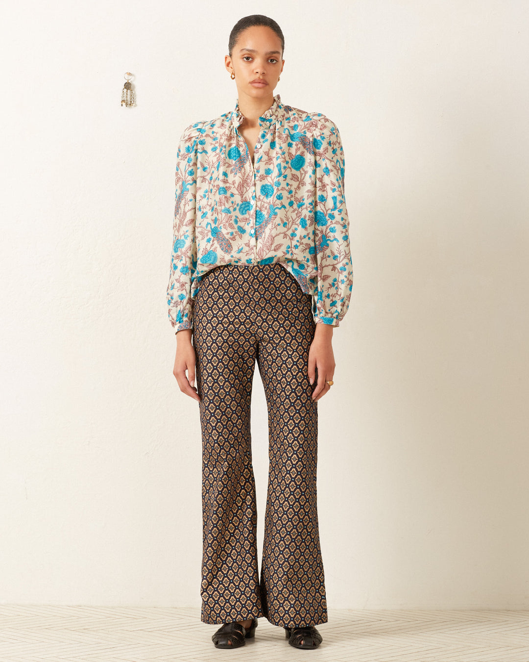 A person stands against a white wall wearing the Annabel Peacock Plume Shirt, a vintage-inspired top with a floral blouse design, complemented by patterned flared pants and black shoes.