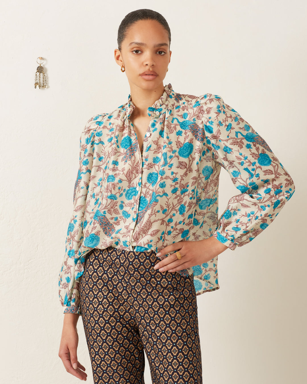 A person wearing the Annabel Peacock Plume Shirt, designed with blue and brown vintage-inspired floral patterns, stands against a plain wall, paired with patterned pants.