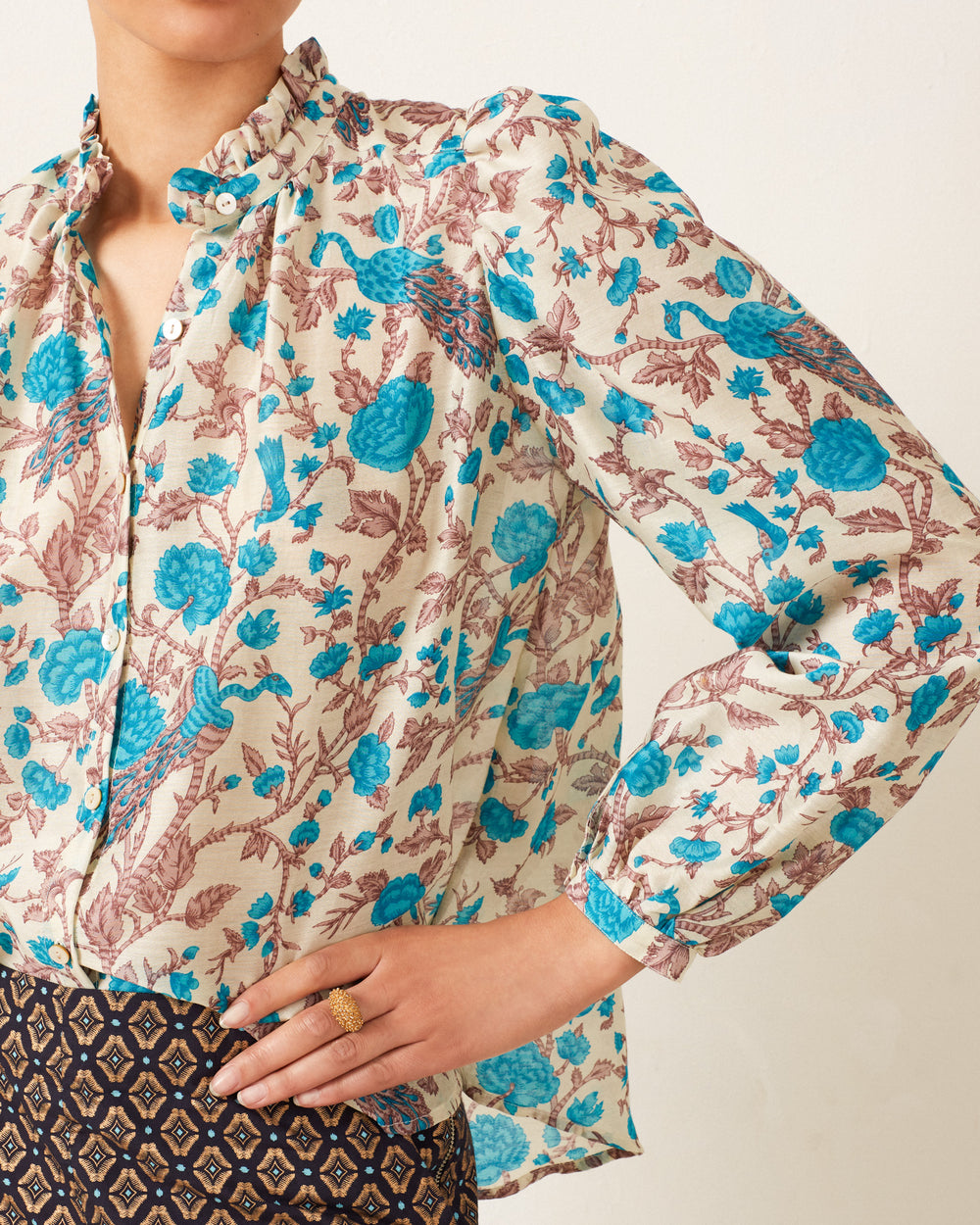 The individual is wearing the Annabel Peacock Plume Shirt, featuring blue and brown botanical patterns, paired with a patterned skirt, and has their hand on their hip.