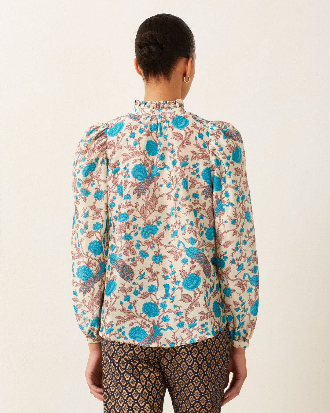 Facing away, the person wears the Annabel Peacock Plume Shirt, featuring vintage-inspired puffed sleeves and paired with patterned pants against a plain background. The cotton-silk fabric blend imparts elegance to the floral blouse, creating a timeless look with a modern twist.