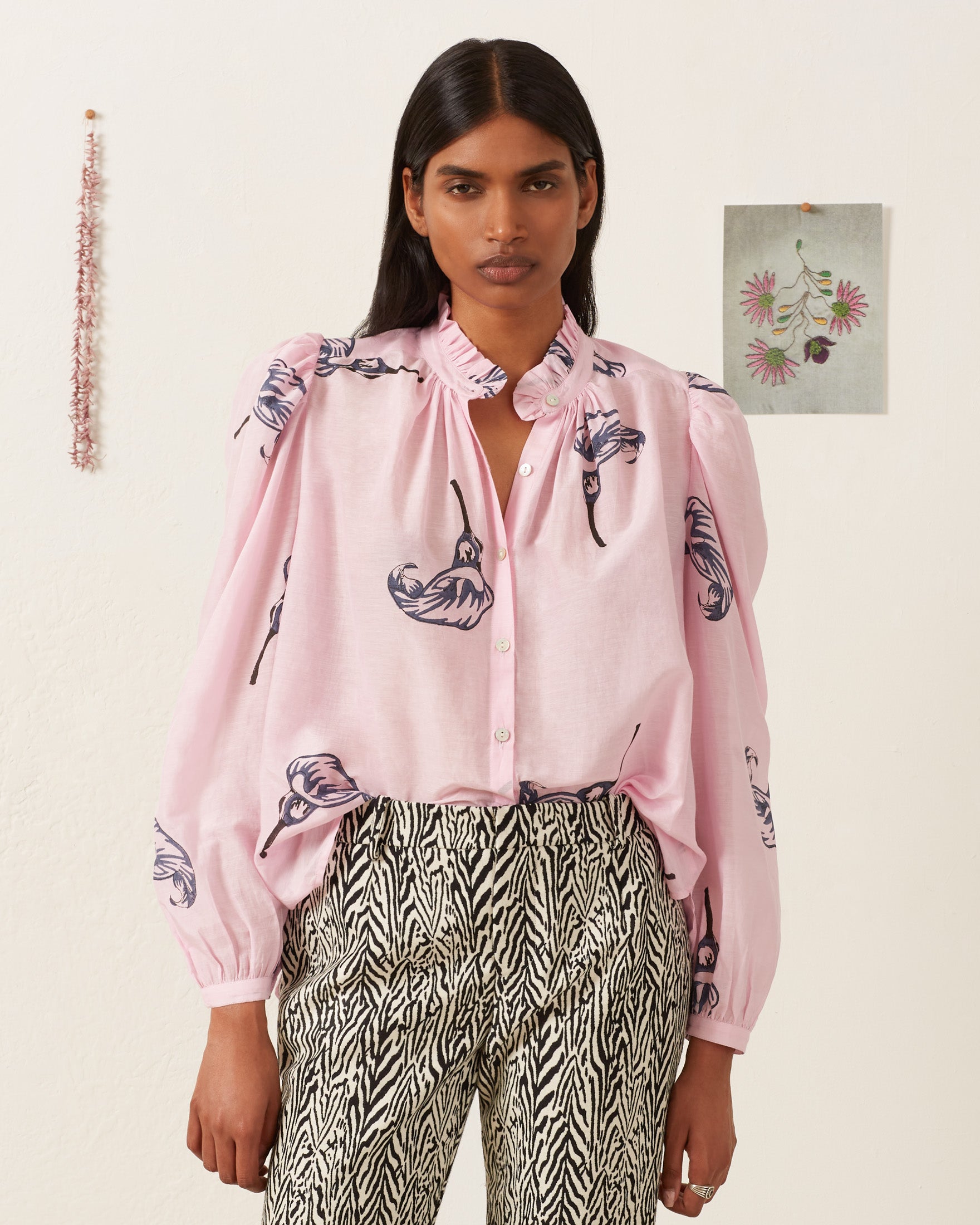 A person showcases the Annabel Pink Calla Lily Shirt, featuring a pink floral blouse with a lily print made from a luxurious cotton-silk blend, standing against a white background with wall decorations.
