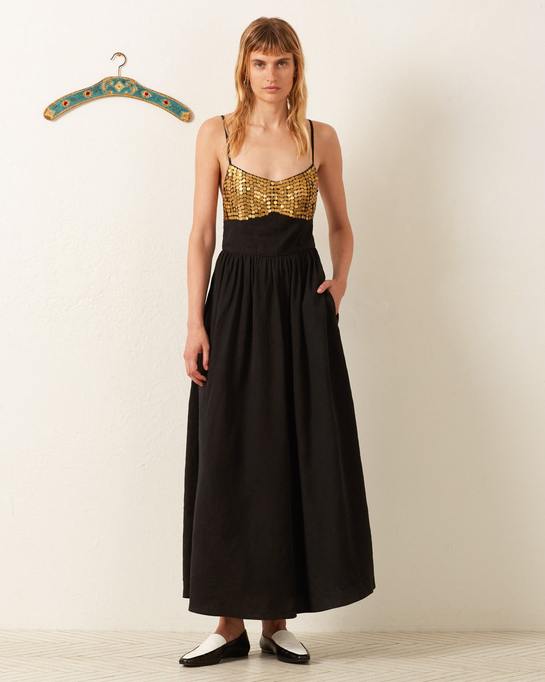 A person wearing an Autumn Blackjack Coin Dress featuring a long black skirt and a gold top with hand-embellished designs stands next to a wall with a decorative hanger.