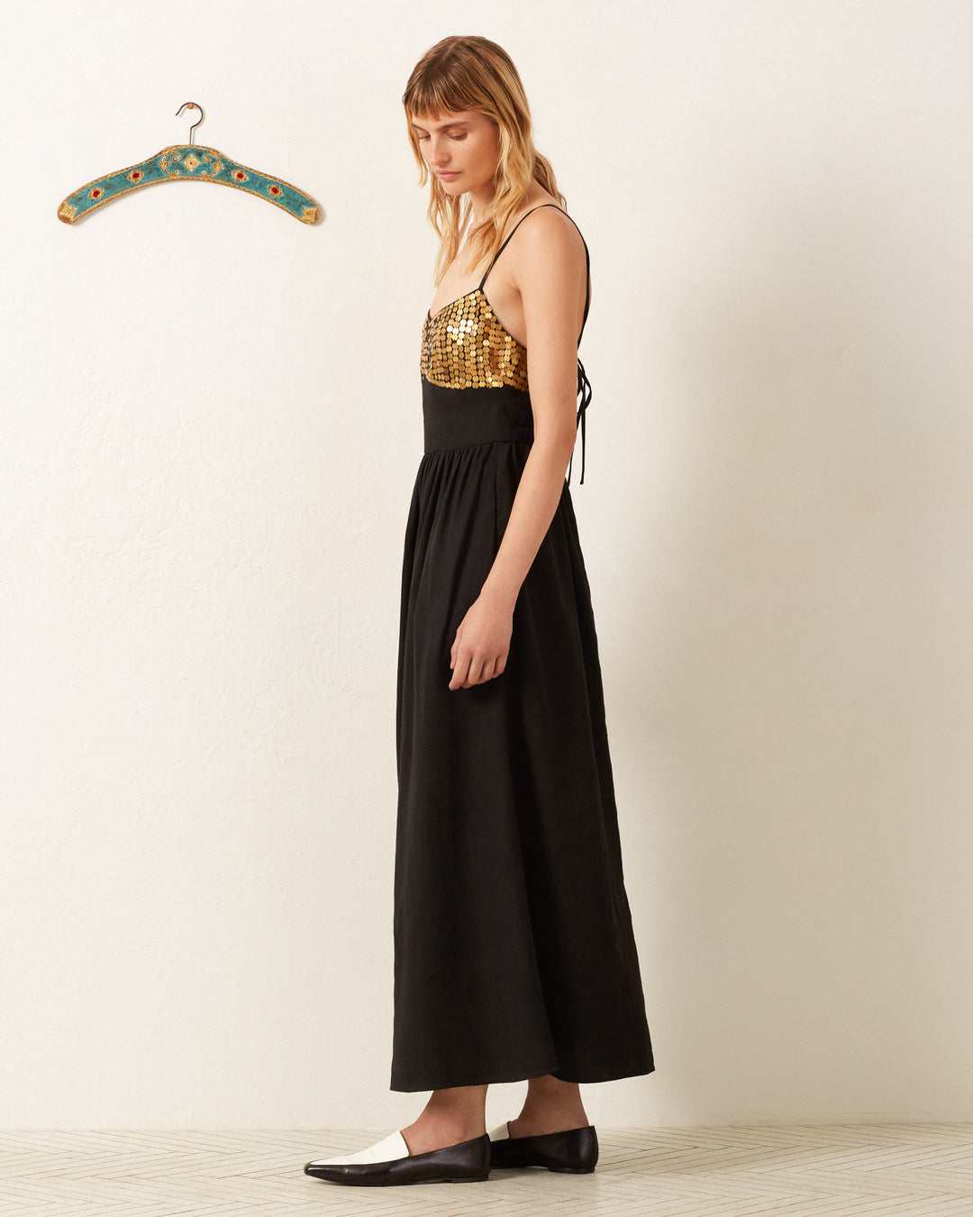 A woman wearing the Autumn Blackjack Coin Dress, characterized by its formal long black skirt and hand-embellished gold metal sequins on the top, stands in profile near a decorative hanger on a white wall, her look complemented by chic black-and-white shoes.