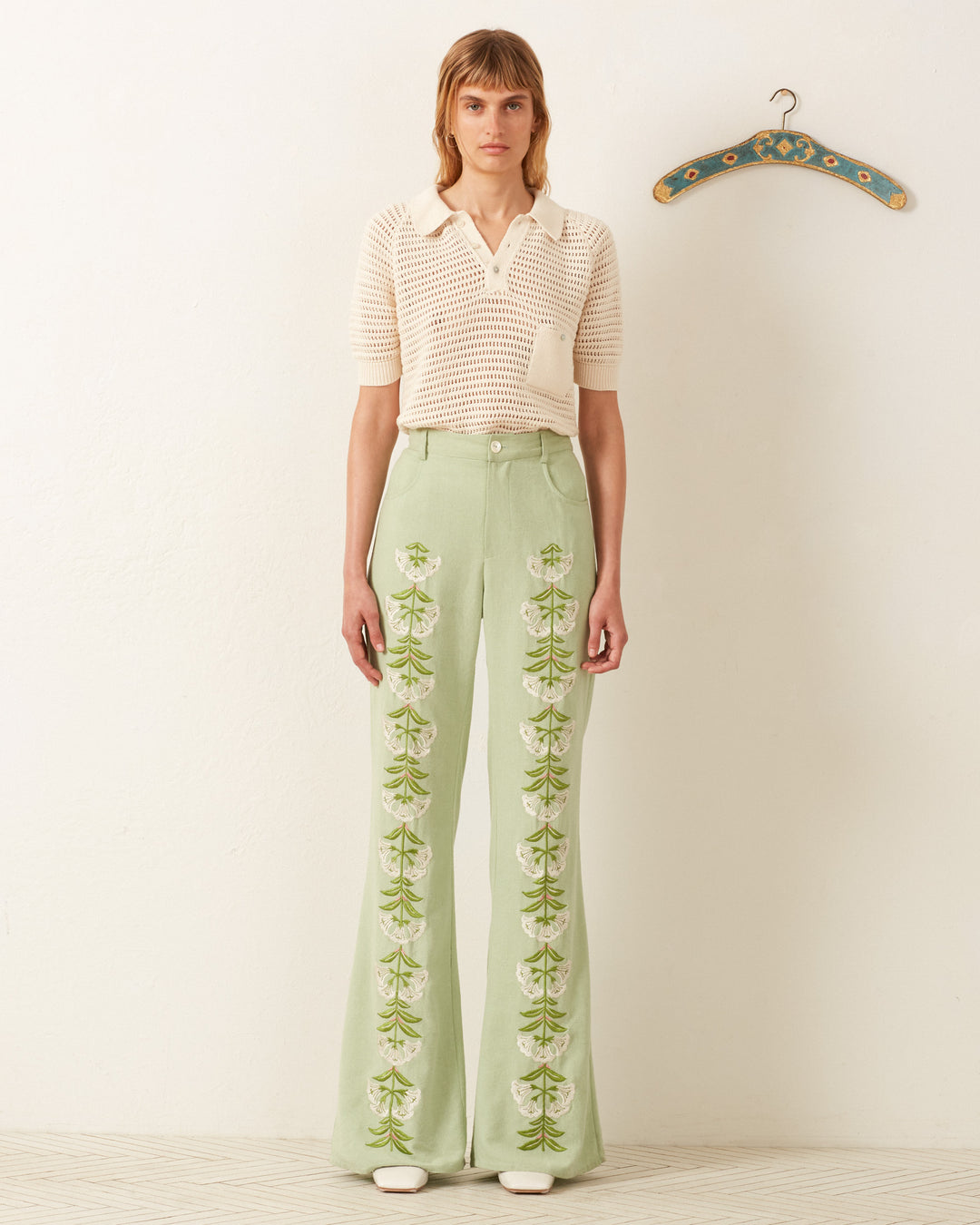 Dressed in a cream short-sleeve sweater and Bunny Trumpet Love Jean with a '70s floral vibe, a person stands in a room with a decorative hanger on the wall, exuding retro charm.