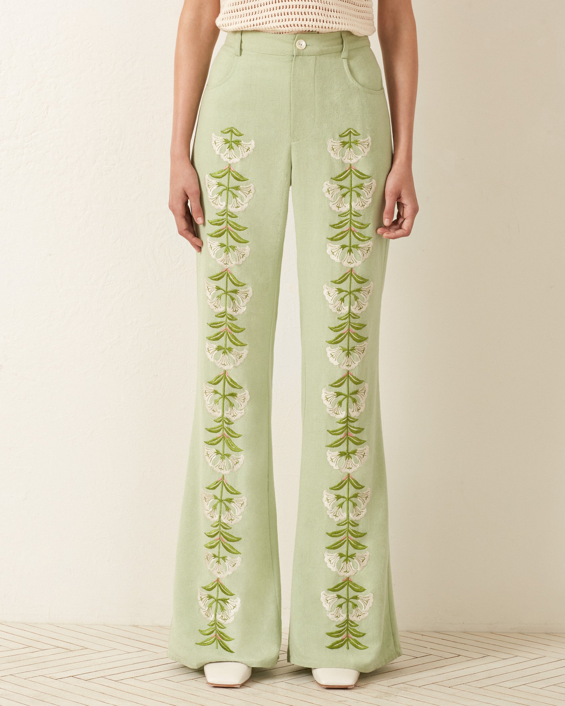 A person in Bunny Trumpet Love Jeans, featuring light green silk with vertical white and green floral embroidery, stands against a neutral background, radiating a '70s vibe.