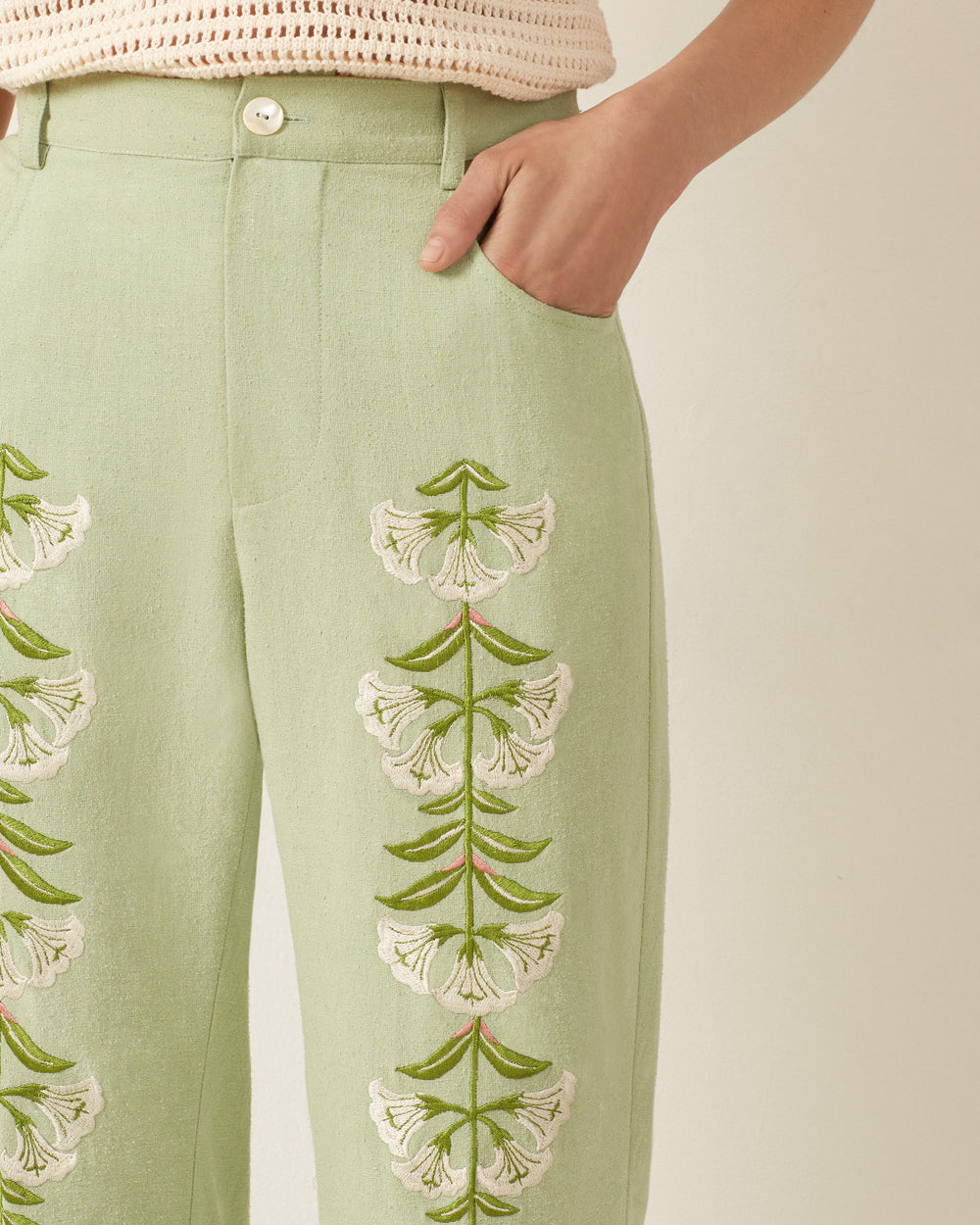 A person wearing Bunny Trumpet Love Jeans in light green silk, featuring a '70s vibe with white and green floral embroidery, right hand in pocket.