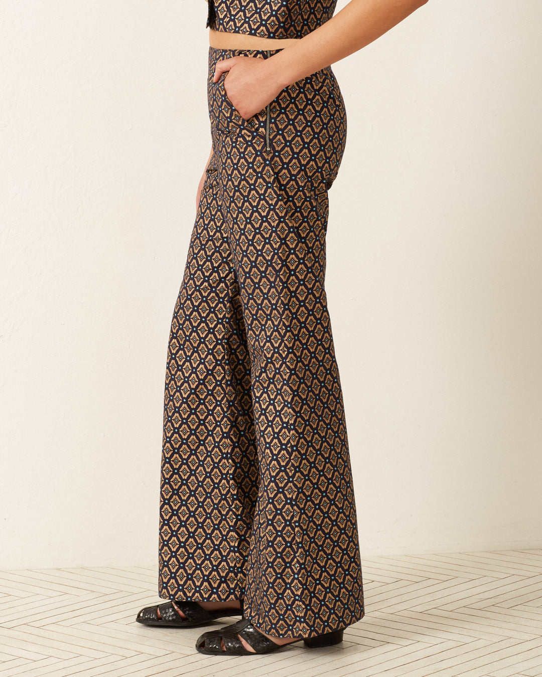 A person wearing the Charlie Graceland Pants, featuring a high-waisted, '70s-style wide-leg design with a dark and light geometric print, stands against a light background. The metallic cotton pants are paired with a matching top, and the person poses with one hand in their pocket.