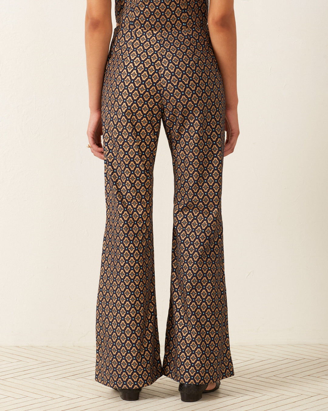 A person wearing the Charlie Graceland Pant, styled with '70s-inspired high-waisted flares adorned with a striking geometric print, stands with their back turned. The floor beneath them is wooden, and the background remains plain.