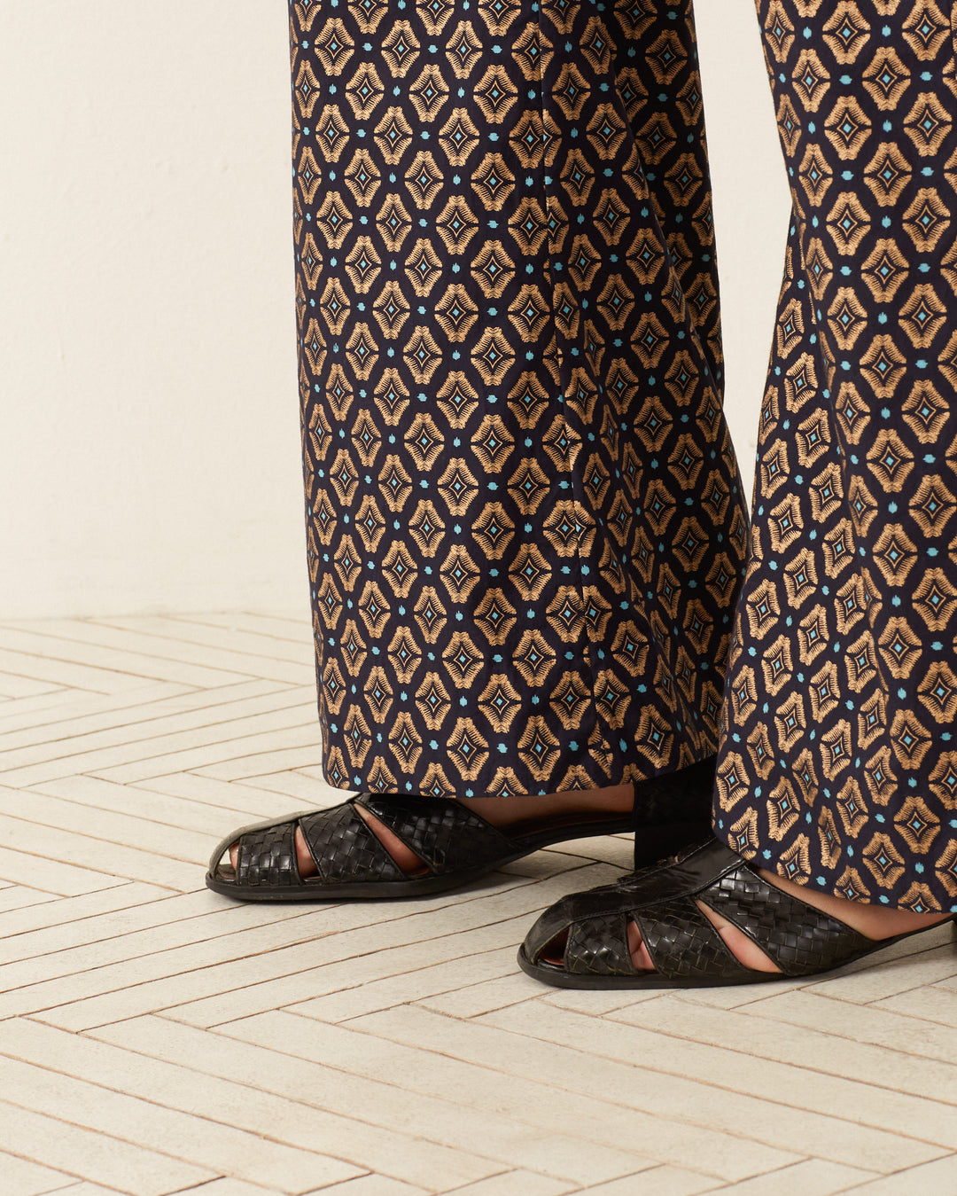 Close-up of a person wearing '70s-style Charlie Graceland Pants featuring a geometric print in brown, black, and blue. They are also wearing black woven sandals. The floor has a light wooden appearance with horizontal lines.