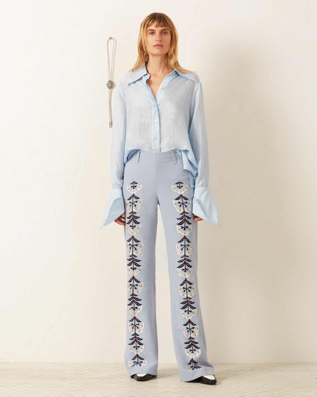 A person wearing the Dolly Bluejay Silk Shirt, a light blue vintage-inspired blouse, along with embroidered trousers stands against a plain wall.