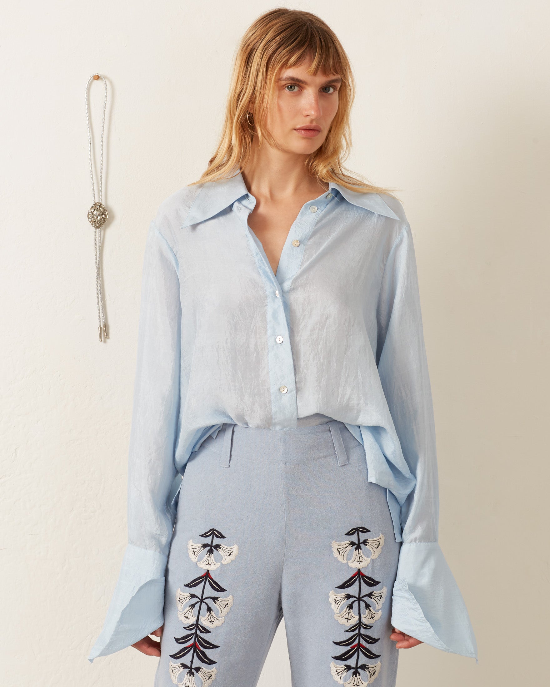 A person with long hair wears the Dolly Bluejay Silk Shirt and high-waisted embroidered pants, creating a vintage-inspired look. A necklace hangs on the wall in the background.