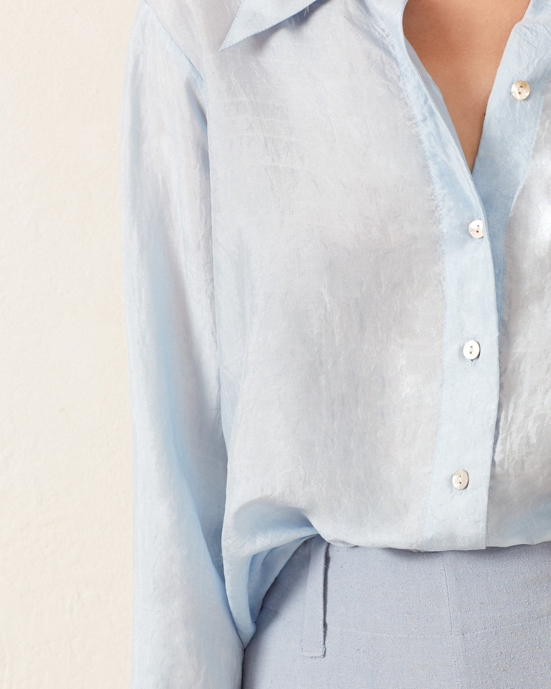 A close-up of a person in a vintage-inspired look, showcasing the Dolly Bluejay Silk Shirt with buttons, partially tucked into matching light blue pants against a beige background.