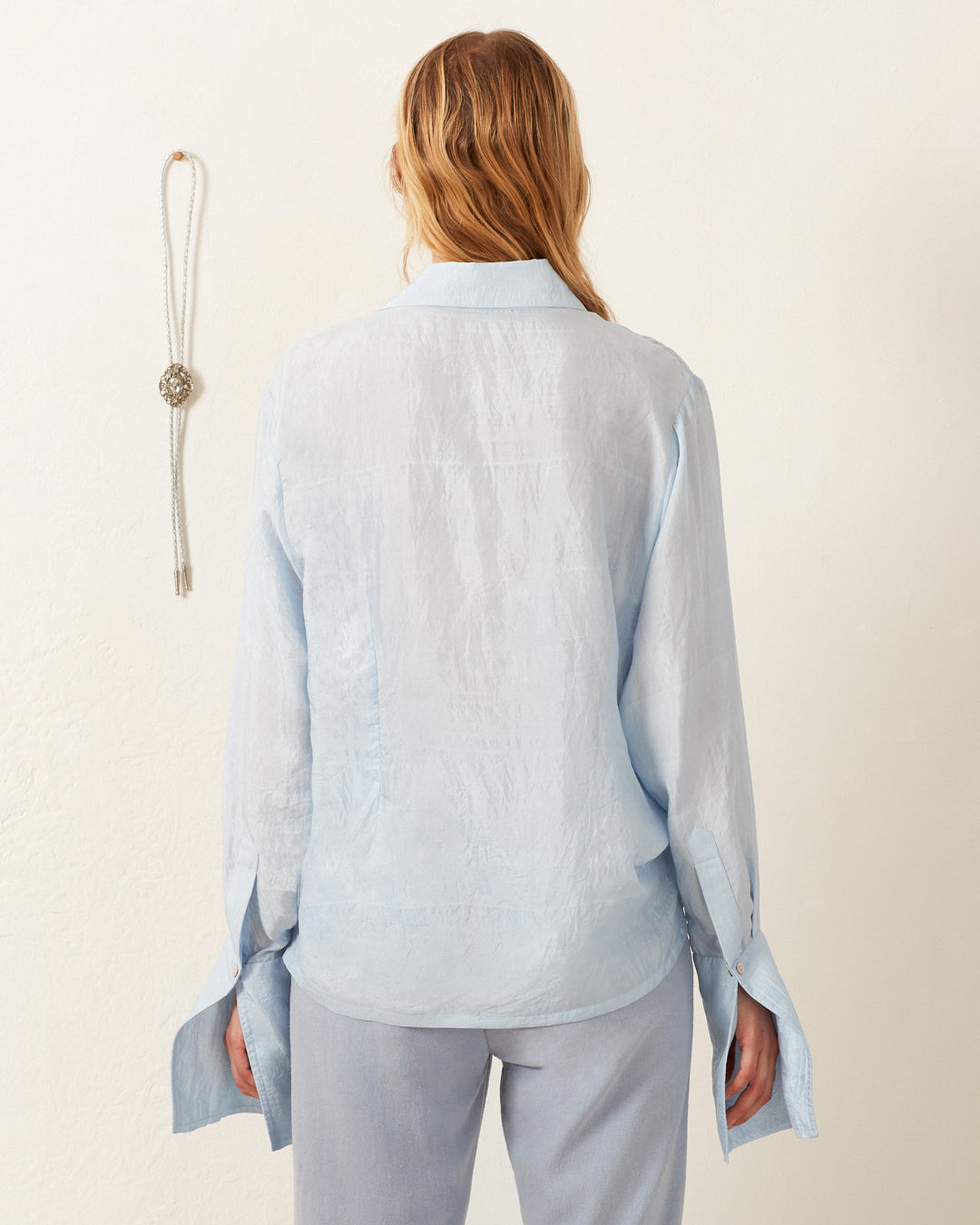 Person facing away in a vintage-inspired look, wearing the Dolly Bluejay Silk Shirt and matching pants with long, loose sleeves. A decorative tassel hangs on the wall.