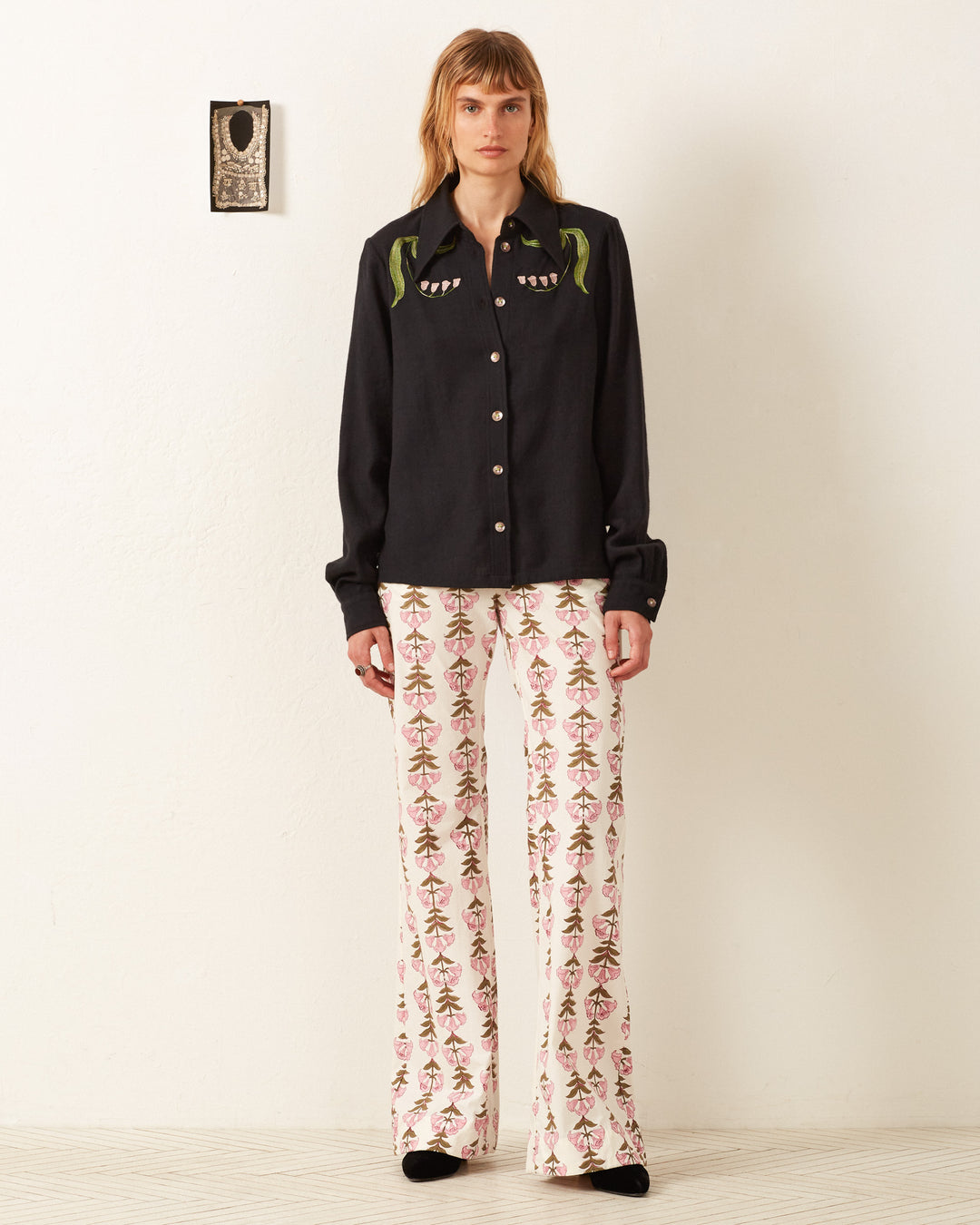 A person stands against a white wall, wearing the Dolly Lily Valley Shirt, a vintage-inspired black button-up with green detailing, paired with white pants featuring a floral pattern.