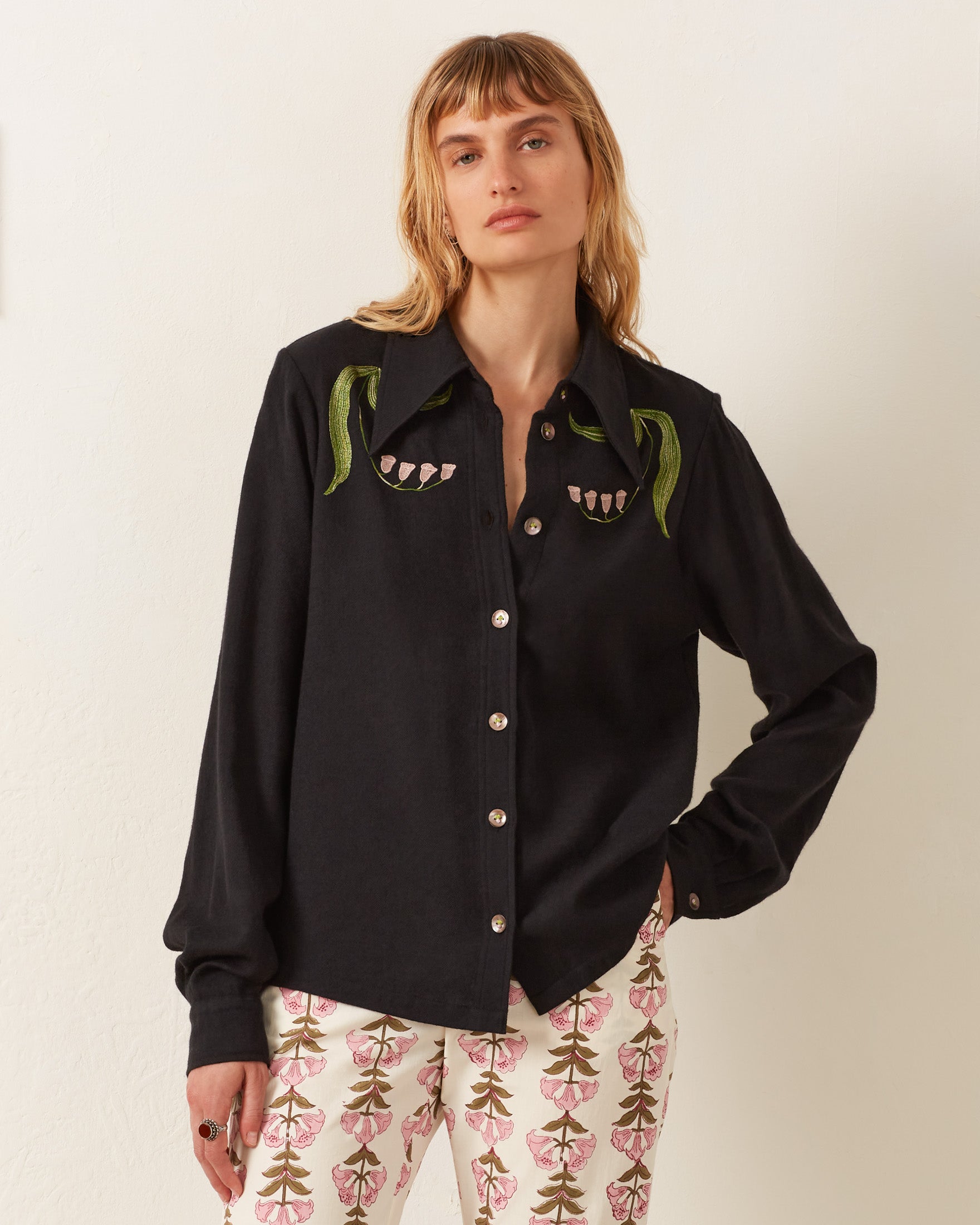 A person wearing the Dolly Lily Valley Shirt, a vintage-inspired black button-up with hand-embroidered plant designs, and white pants featuring a floral pattern stands against a plain background.