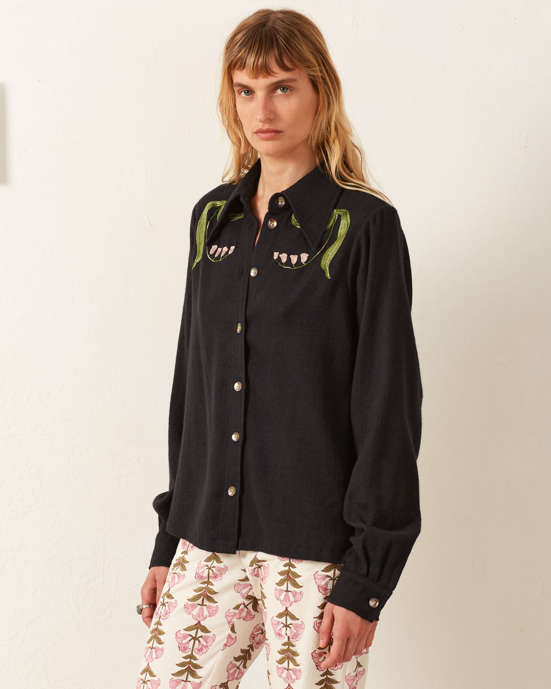 A person wearing the Dolly Lily Valley Shirt, a vintage-inspired black shirt with hand-embroidered details, along with patterned pants, stands against a plain background.