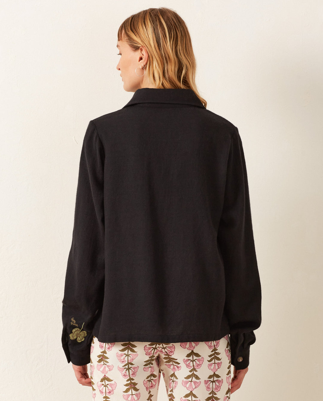 A person with shoulder-length hair is wearing a vintage-inspired Dolly Lily Valley Shirt in black and patterned pants, standing with their back to the camera against a plain white wall.