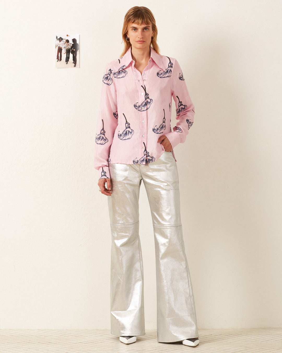 A person wearing a Dolly Pink Calla Lily Shirt and shiny silver pants stands in a room with a small photo on the wall.