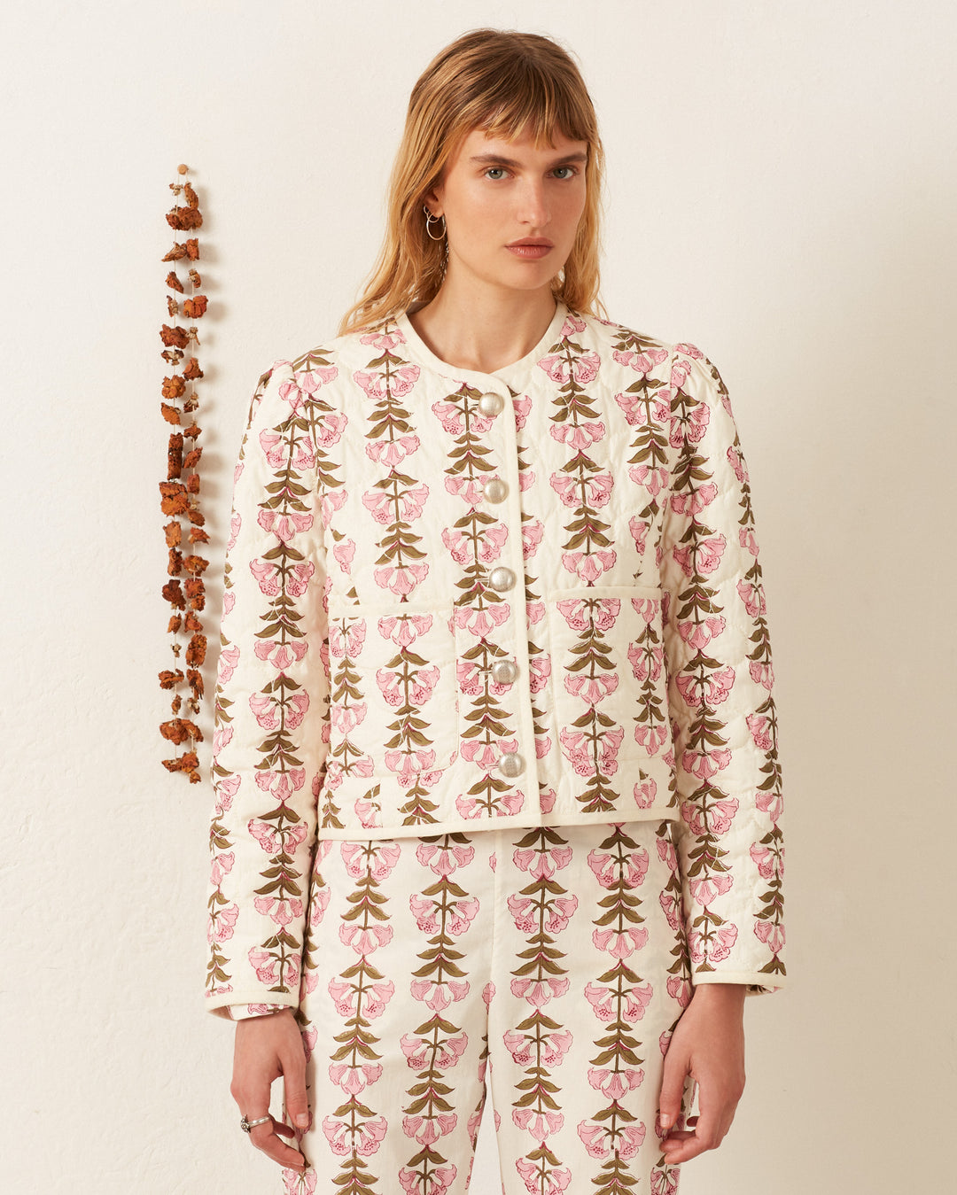 Dressed in a Dorian Trumpet Lily Jacket and coordinating pants, an individual stands before a simple backdrop enhanced by a vertical line of dried flowers.