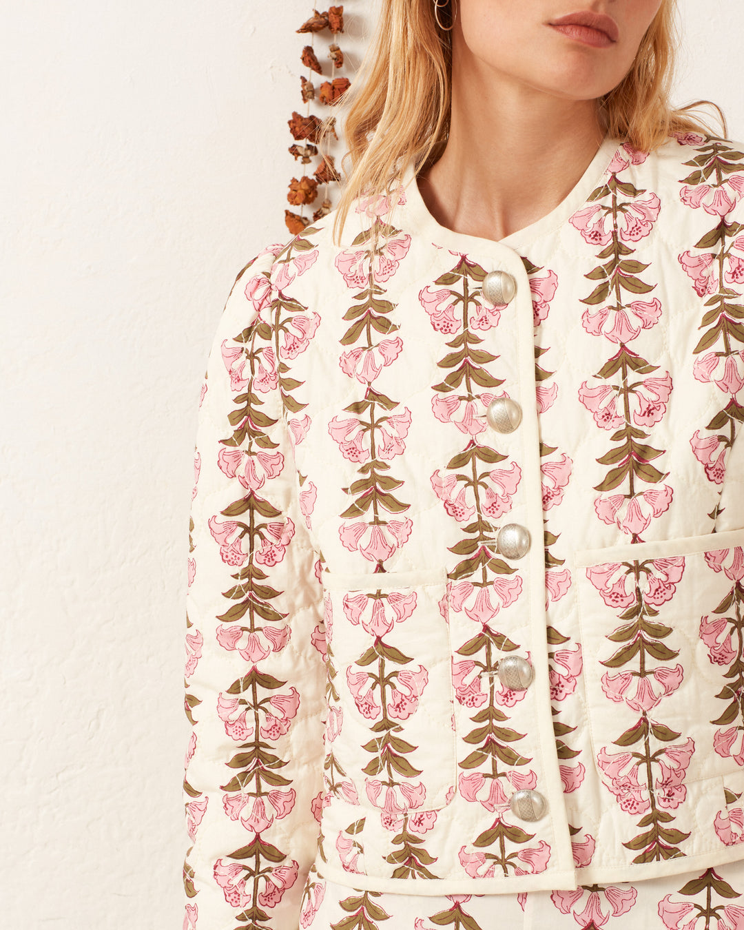 Against a white wall, a woman wears the Dorian Trumpet Lily Jacket, showcasing its cream color and pink-and-green floral pattern, echoing hand-quilted artistry.