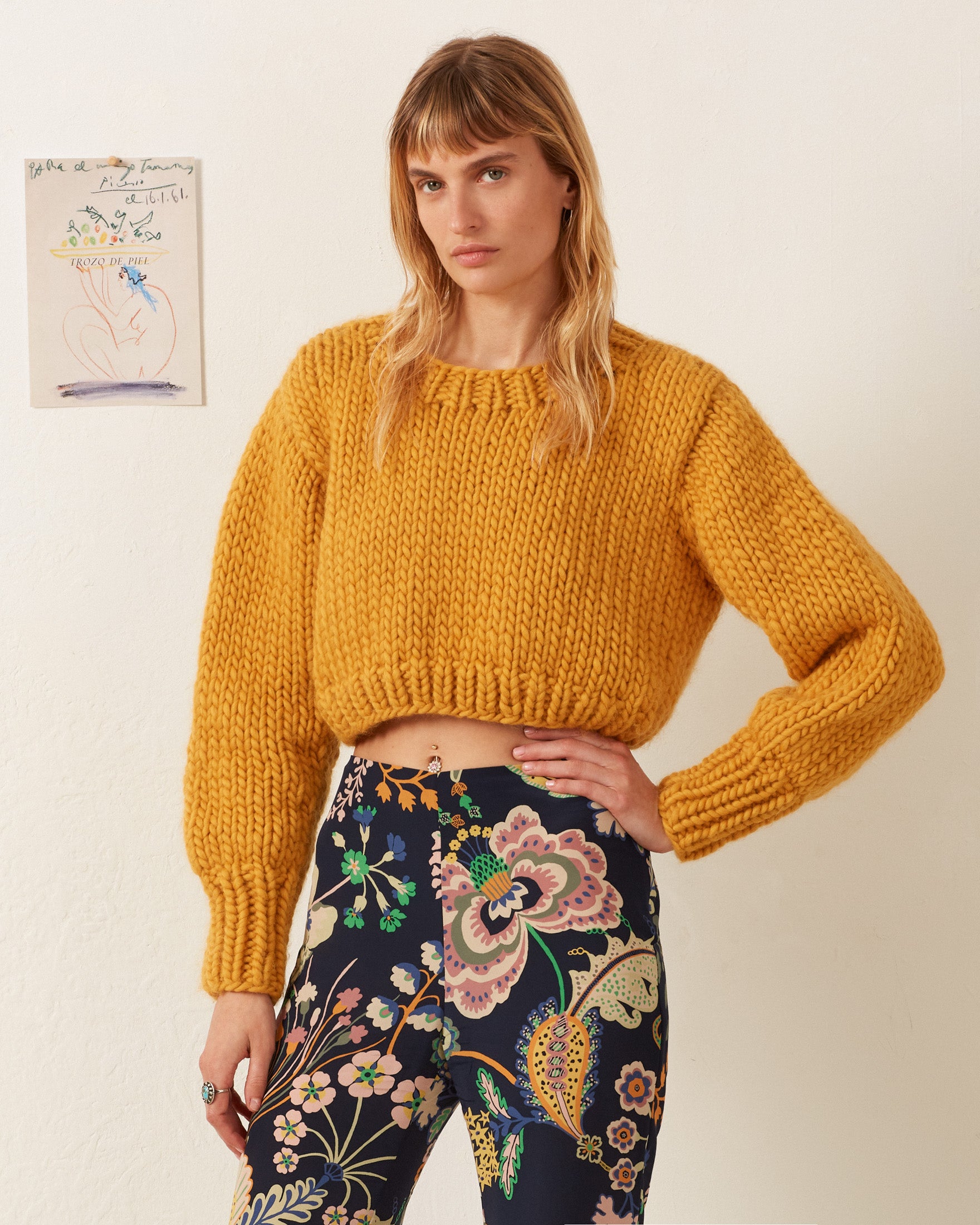 A person stands against a wall, their Ella Mango Knit Sweater perfectly complementing the vibrant floral pants. An intricate drawing on the wall adds artistic flair to the scene.