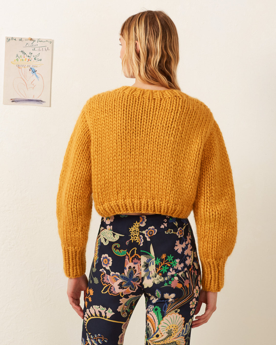 A long-haired individual wears an Ella Mango Knit Sweater with patterned pants, standing in front of a beige wall with small artwork taped to it.