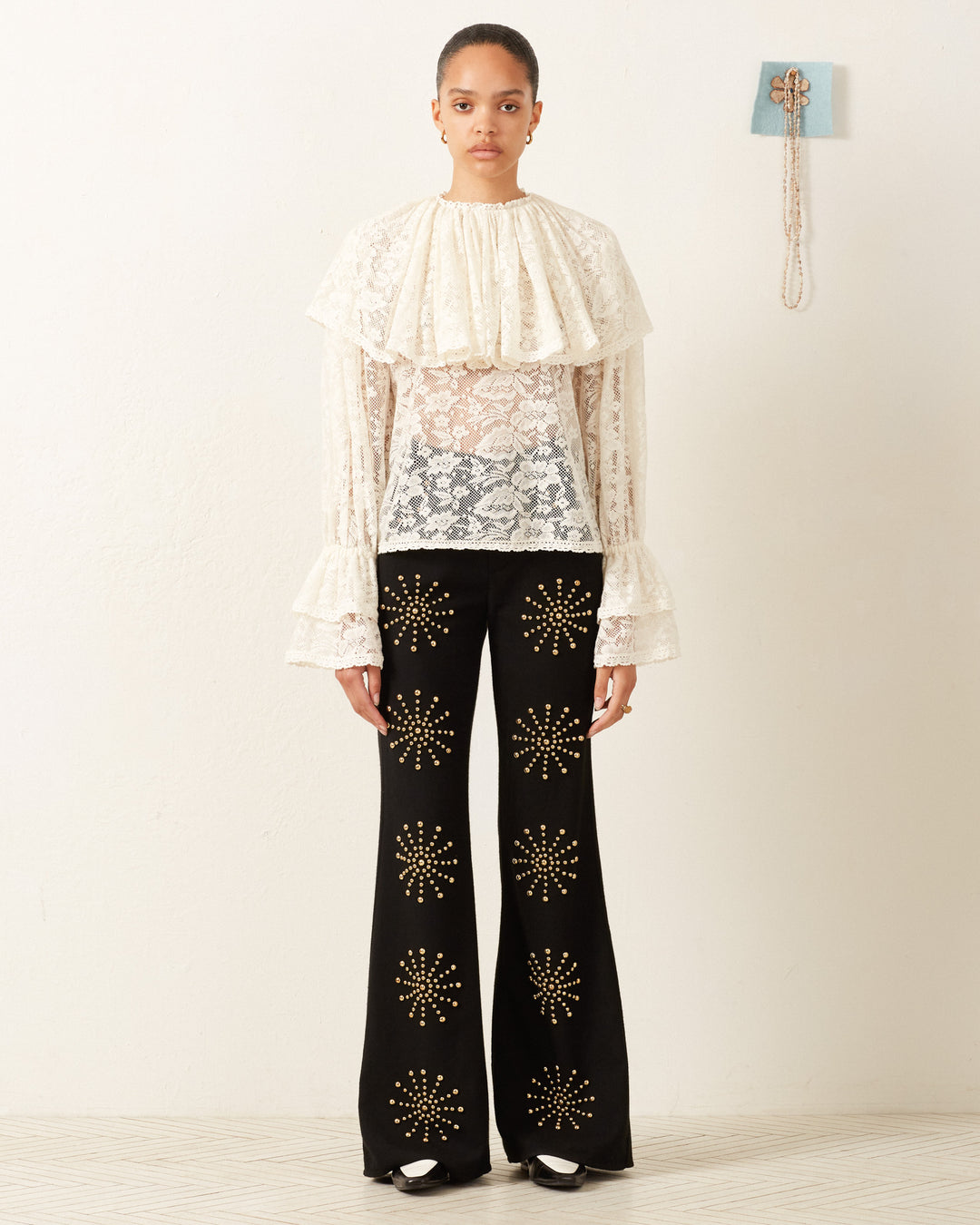 A person wearing the Eloise Magnolia Lace Top, characterized by its vintage feel and ruffled sleeves, paired with black pants adorned with floral patterns, stands against a plain wall. A necklace hangs on the wall to the right.