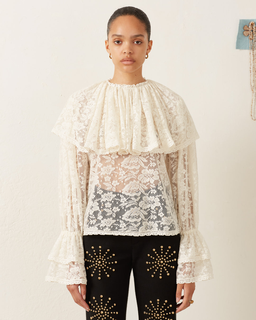 A person wearing an Eloise Magnolia Lace Top with sheer white floral lace and ruffled sleeves, paired with black pants featuring gold star patterns, stands against a plain white background, exuding a vintage feel.