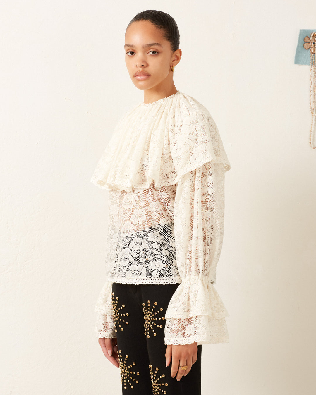 A person wearing the Eloise Magnolia Lace Top, which features a vintage-feel white floral lace with ruffled sleeves, along with black pants adorned with gold embroidery, stands against a plain background.