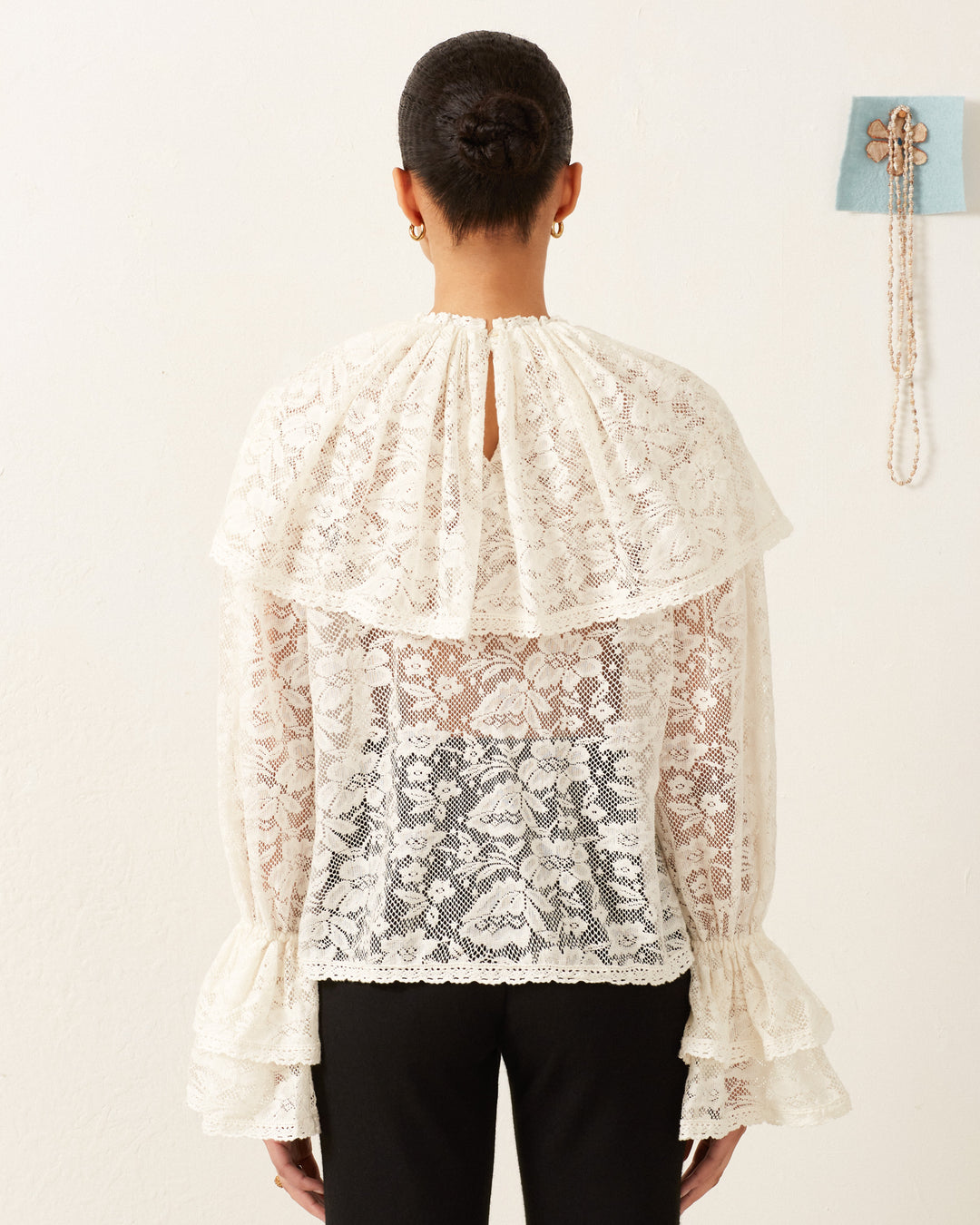 A person is shown from behind wearing the Eloise Magnolia Lace Top, characterized by its vintage-feel white lace and ruffled sleeves. Their hair is styled in a bun, and they are facing a white wall decorated with art that includes a beaded accessory.