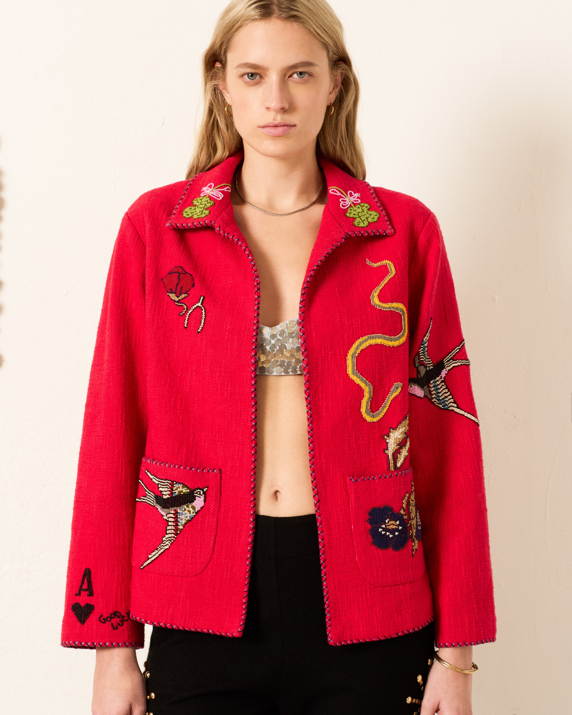A woman wears the Francis Crimson Tattoo Jacket, a red, hand-beaded embroidered piece with bird and floral designs, exuding vintage charm. She elegantly pairs it with black pants against a plain, light-colored background.