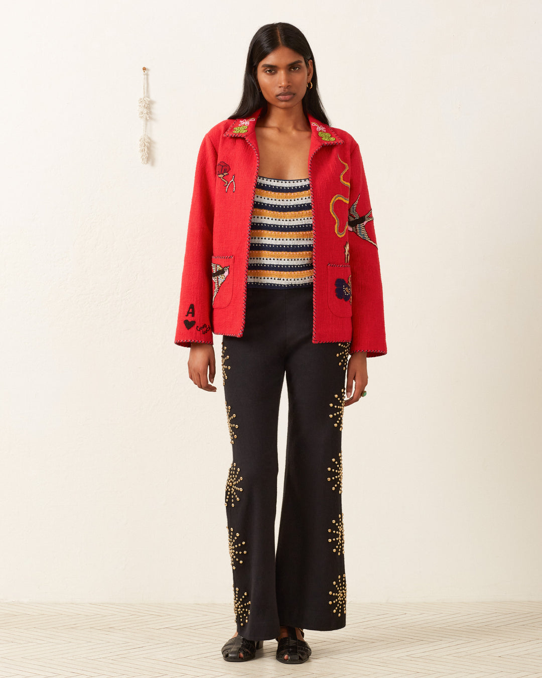 A person wearing the Francis Crimson Tattoo Jacket with hand-beaded design details and a striped top pairs perfectly with black pants featuring gold embellishments, exuding a vintage feel against the plain background.