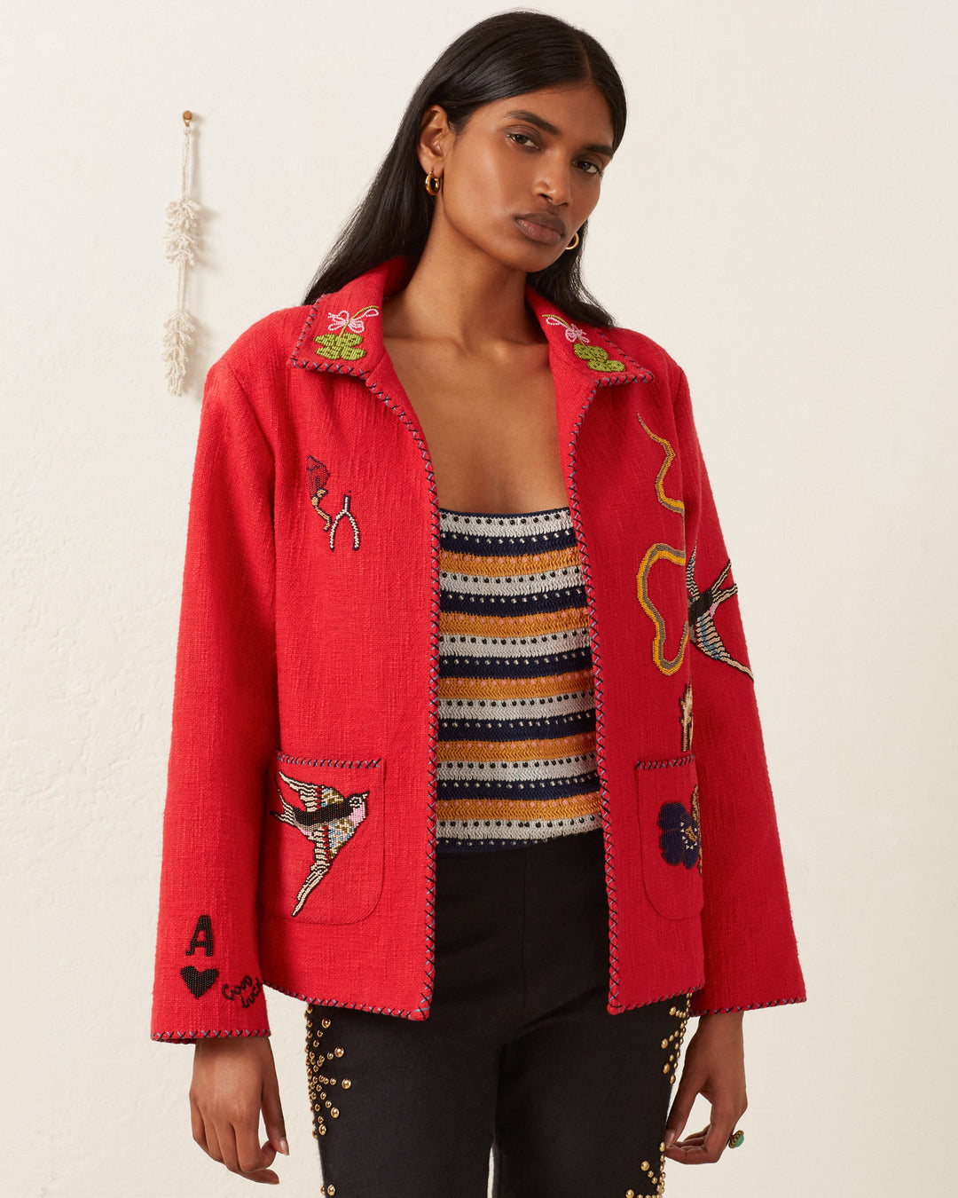 A person wearing the Francis Crimson Tattoo Jacket, a hand-beaded red piece, over a striped top, stands against a plain background exuding a vintage feel.