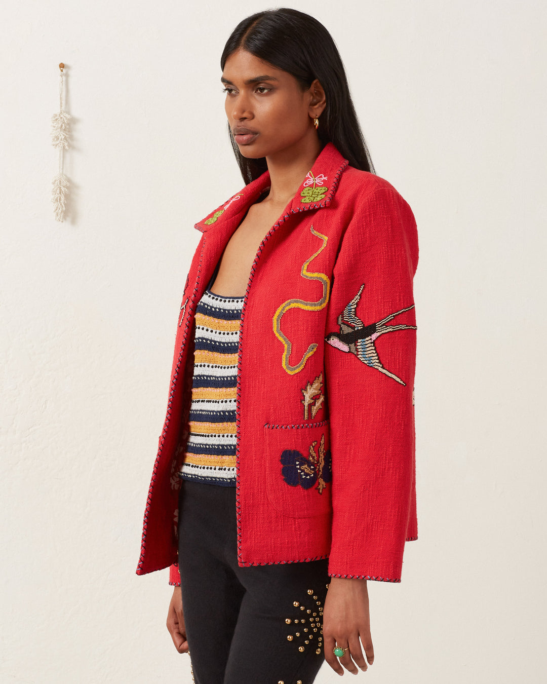 Sporting the Francis Crimson Tattoo Jacket, a person stands against a plain background, conveying a vintage feel with its red embroidered bird and butterfly designs over a striped top, enhanced by intricate hand-beaded details.