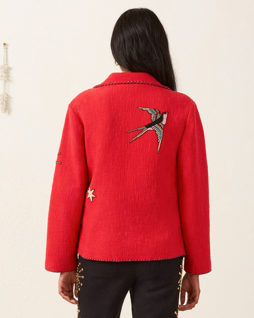 A person with long hair wearing the Francis Crimson Tattoo Jacket, which exudes a vintage vibe and showcases swallow embroidery on the back, stands facing away in a minimalist setting.