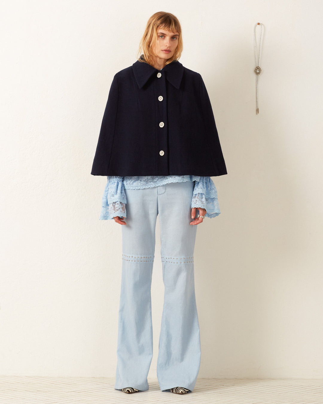 A person wearing the Kensington Midnight Wool Cape, which features a pointed collar, over a blue lace blouse and light blue wide-leg pants stands against a plain white wall. A small wall hanging is visible to the right.