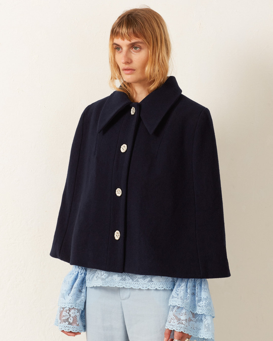 Against a light background, a person wears the Kensington Midnight Wool Cape, featuring large buttons and a pointed collar, over a blue lace-sleeve top and light blue pants.