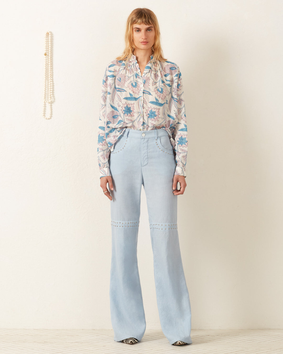 A person stands against a plain white wall, wearing the Kiki Blue Valentine Shirt and light blue, high-waisted, wide-leg jeans. A string of pearls hangs on the wall to the left. The floor is light-colored and textured.