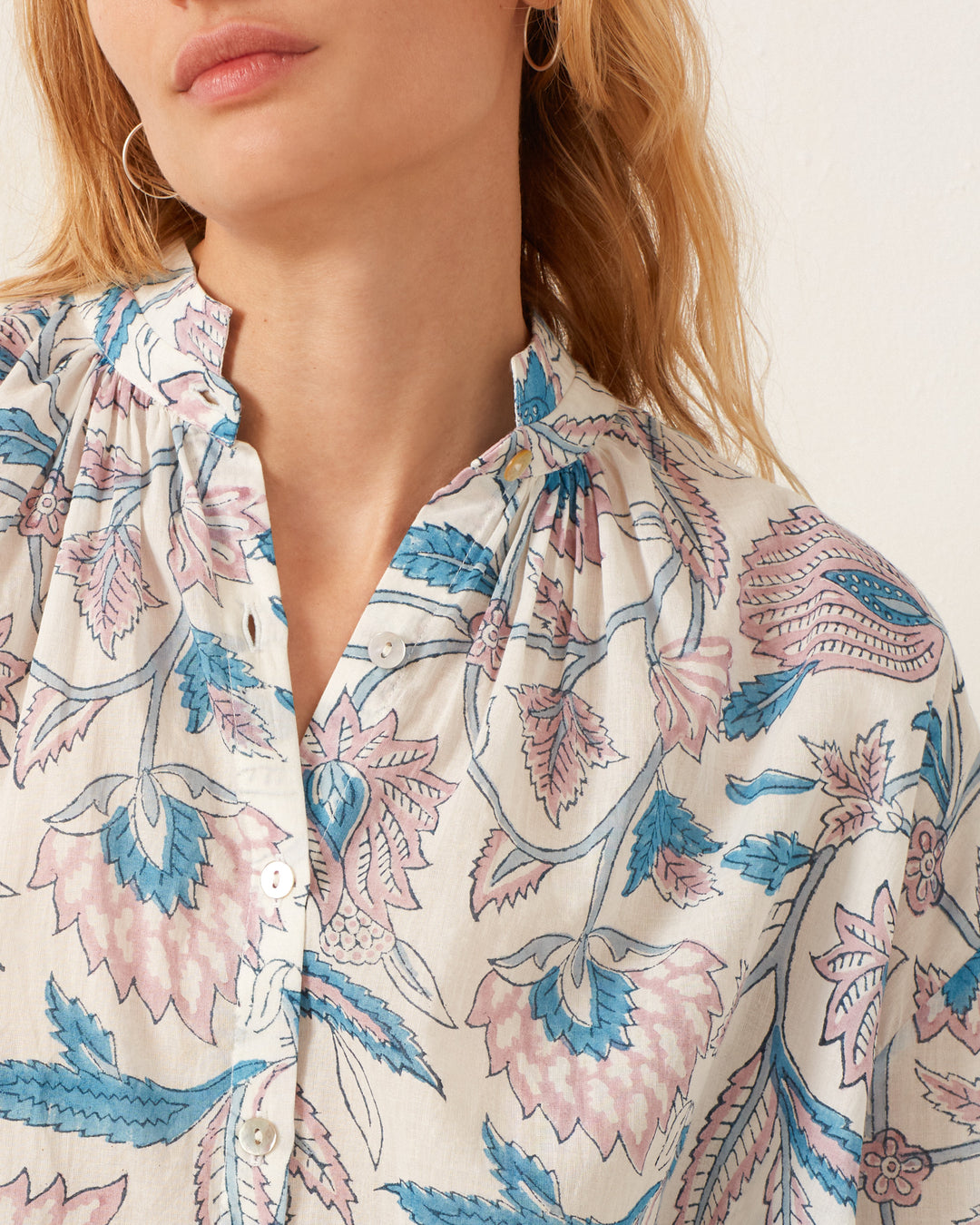 Wearing the Kiki Blue Valentine Shirt, featuring hand-block printed pastel pink and blue floral designs, buttons, and a gathered collar in cotton voile. Their blonde hair partially shows against an off-white wall.