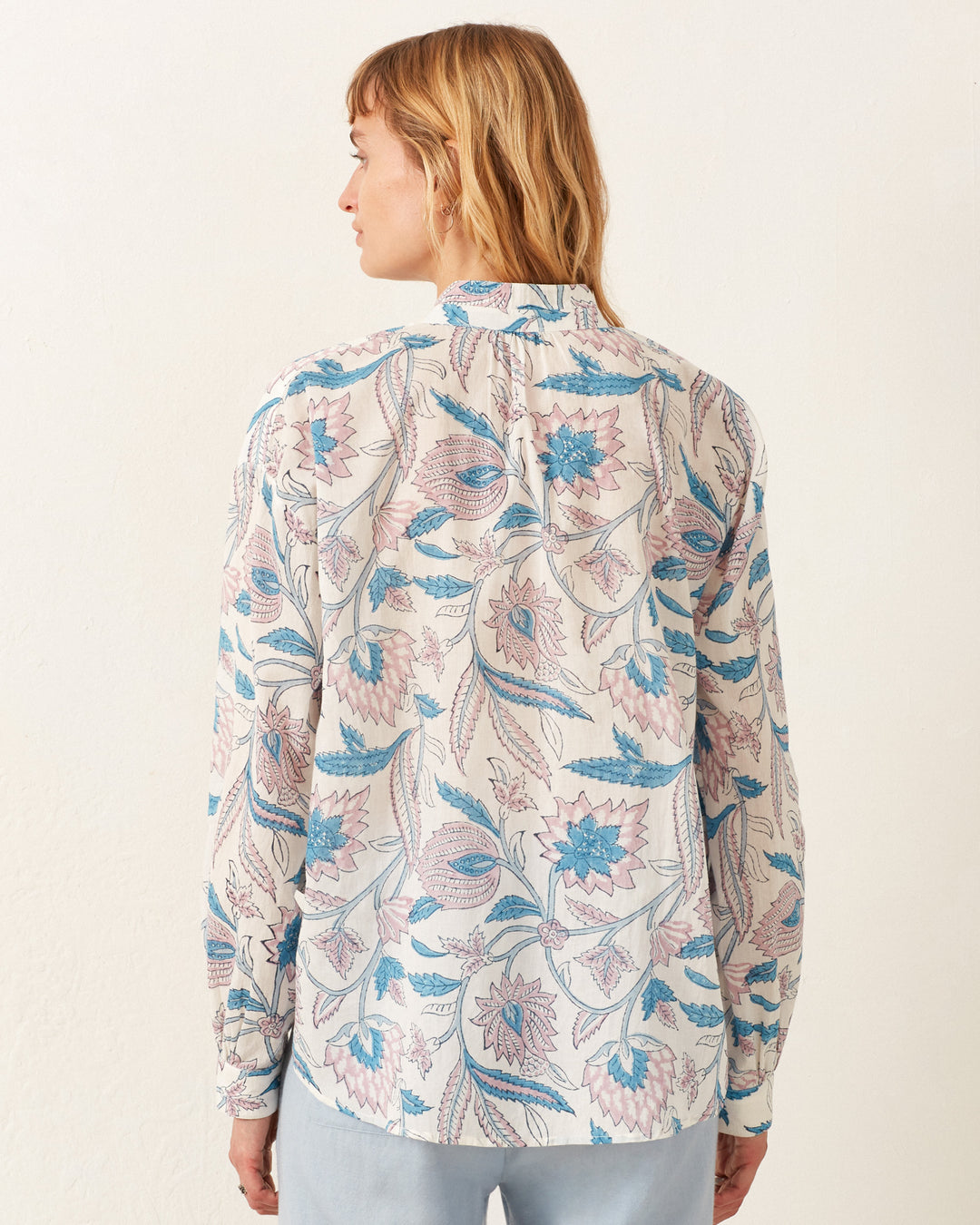 A person with shoulder-length hair faces away from the camera, wearing the Kiki Blue Valentine Shirt. This long-sleeved, button-up floral shirt is made of hand-block printed cotton voile in shades of blue and pink against a plain, light-colored wall.