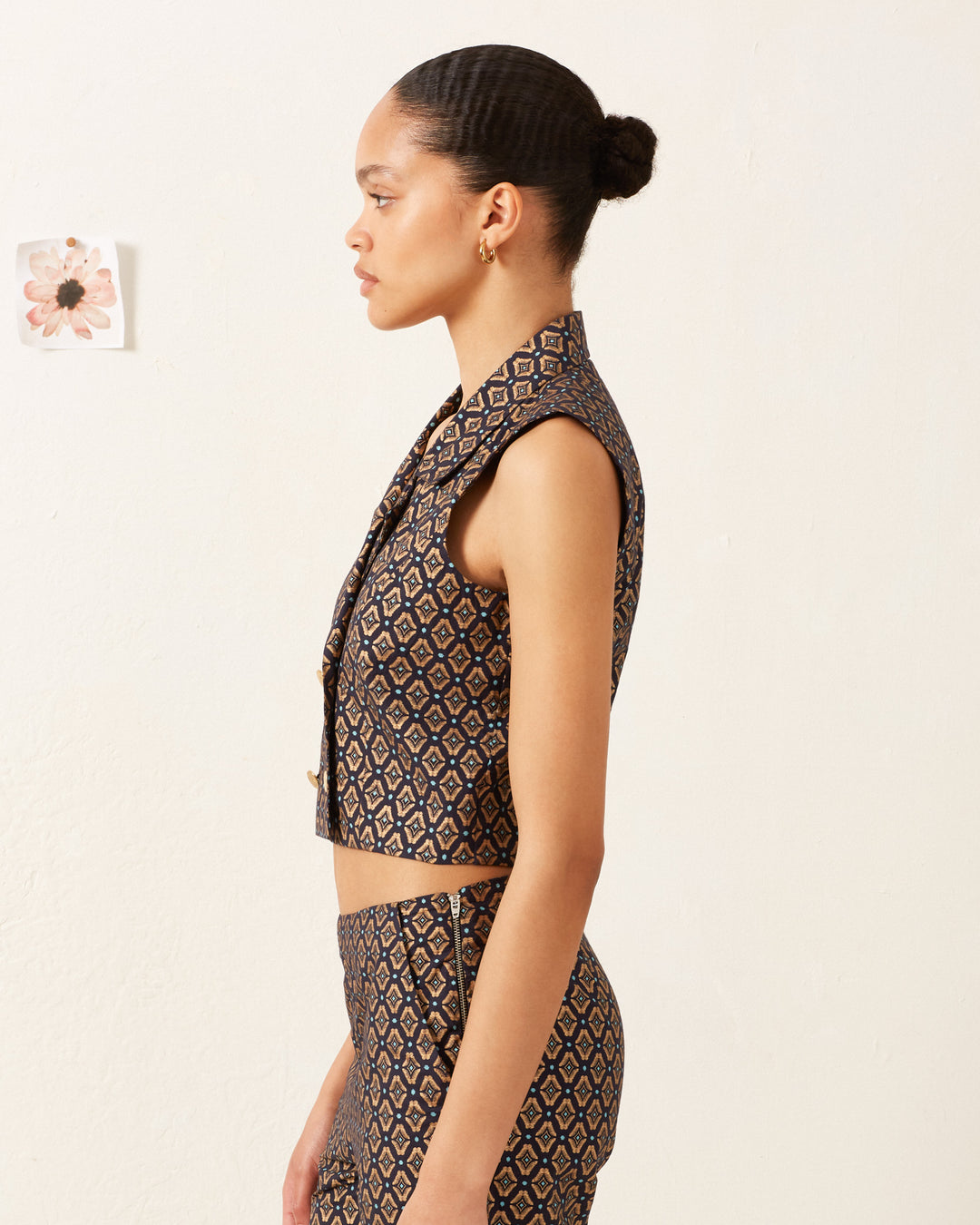 A person with their hair in a bun is wearing a metallic double-breasted Leo Graceland Vest made of cotton, along with matching patterned pants, standing in profile against a light background. St. Christopher buttons glint subtly on the vest, while a small floral picture adorns the wall behind them.