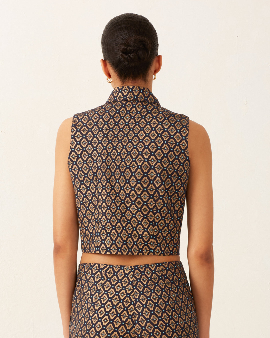 The back view showcases an individual dressed in the Leo Graceland Vest, a sleeveless top with a high collar and matching pants. This ensemble is adorned with a diamond-shaped pattern in shades of blue and beige, highlighted by a subtle gold foil geometric print. The person's hair is elegantly styled in a bun against a plain white backdrop.