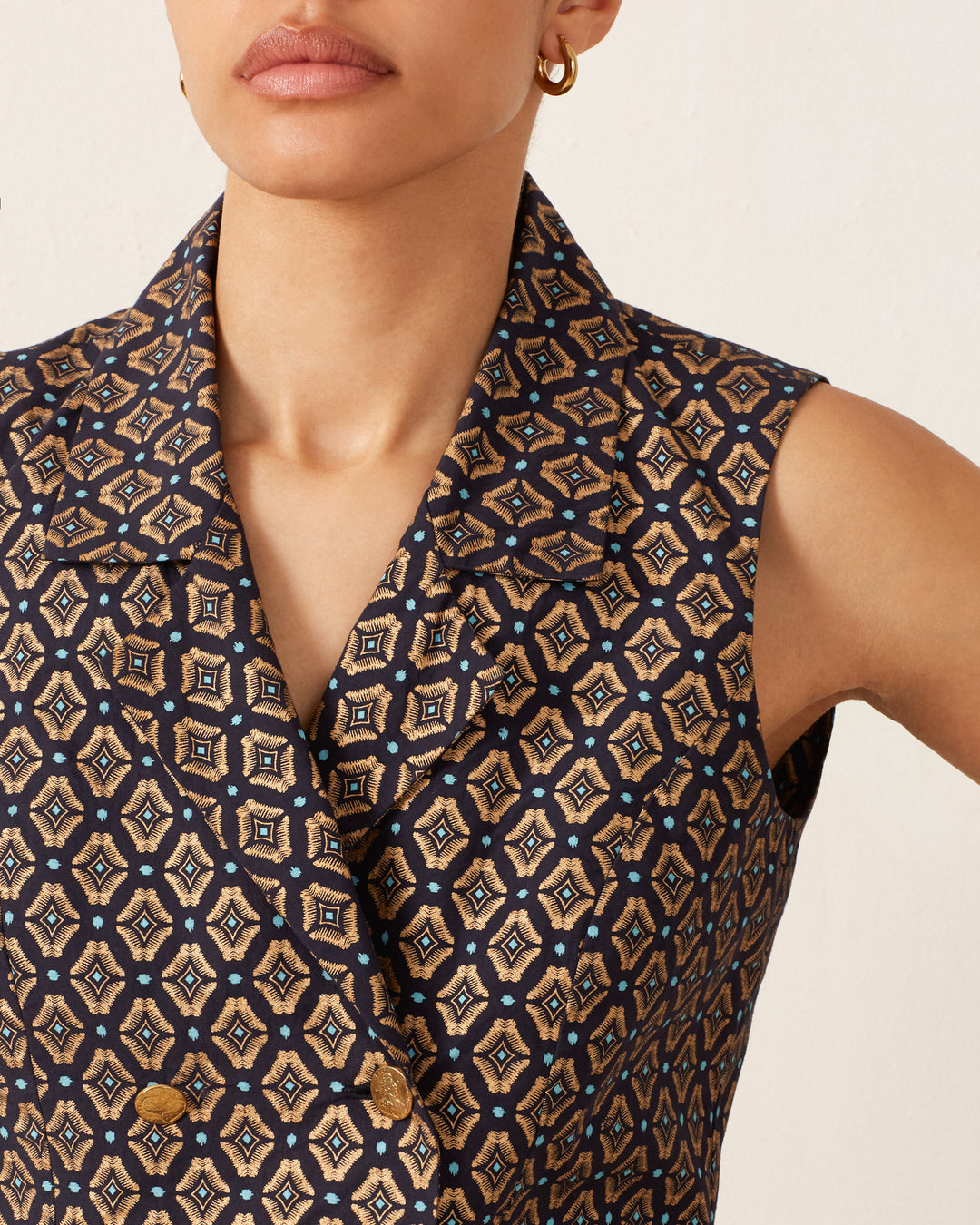 A person wearing the Leo Graceland Vest, a sleeveless top in black and blue with a gold foil geometric pattern. The vest is designed like a metallic double-breasted cotton piece, featuring a collar and two St. Christopher gold buttons. They complete the look with small hoop earrings.