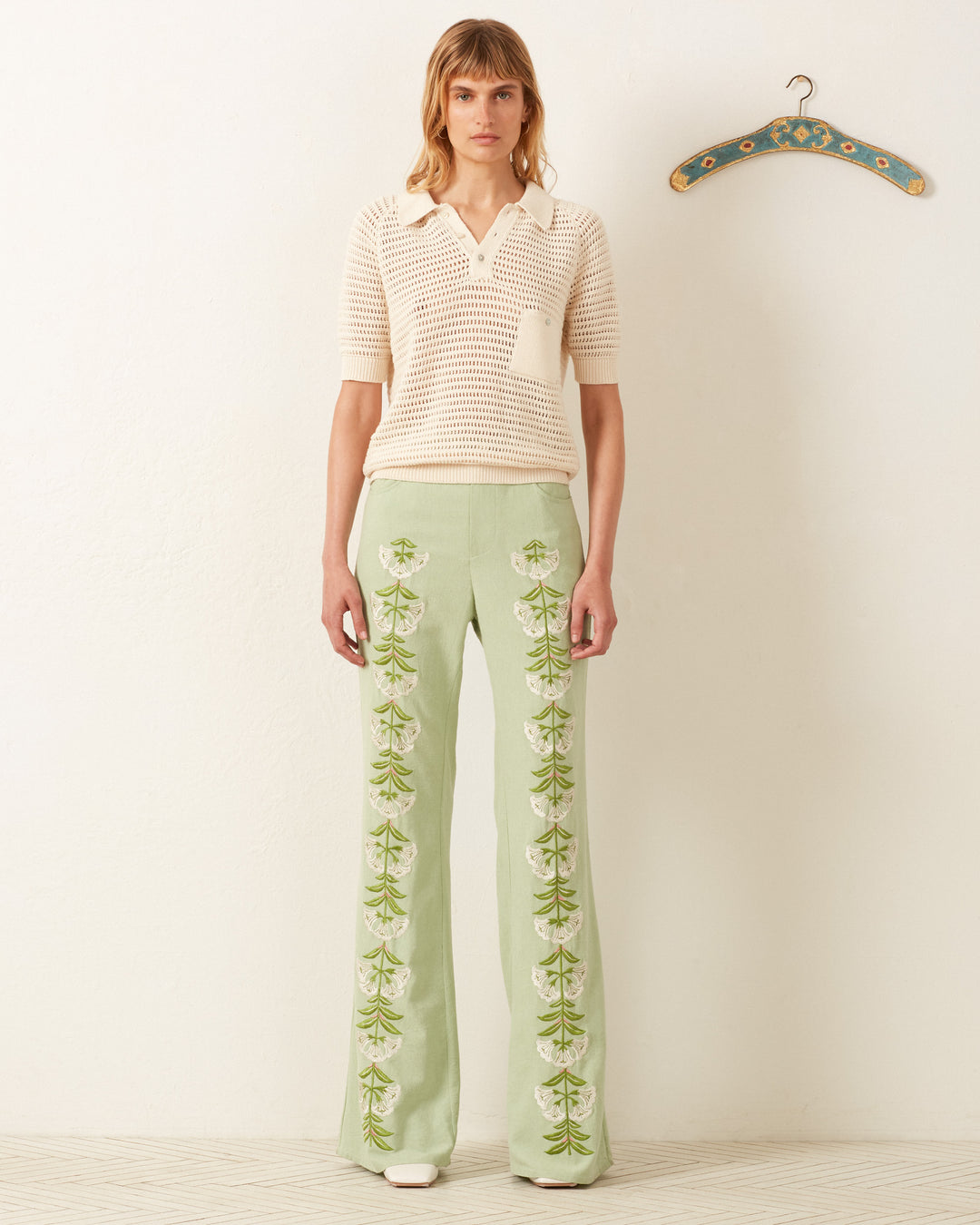 A person stands against a neutral wall wearing the vintage-inspired Lessi Oyster Crochet Top paired with light green pants featuring floral embroidery, exuding Katharine Hepburn's style. A decorative hanger is mounted on the wall.