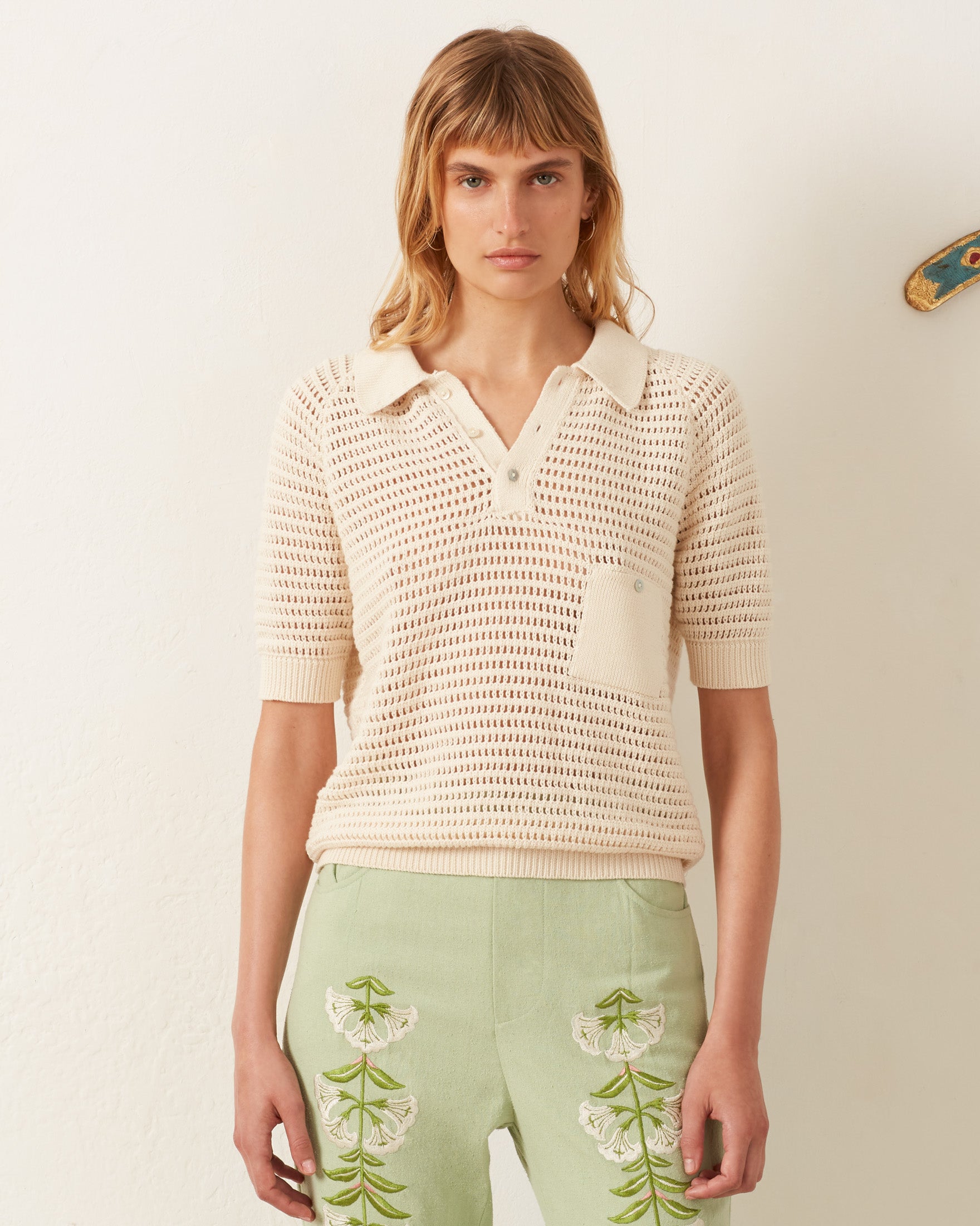 A person with shoulder-length hair wears a vintage-inspired Lessi Oyster Crochet Top and light green pants with floral embroidery, reminiscent of Katharine Hepburn's style, standing against a plain white background.