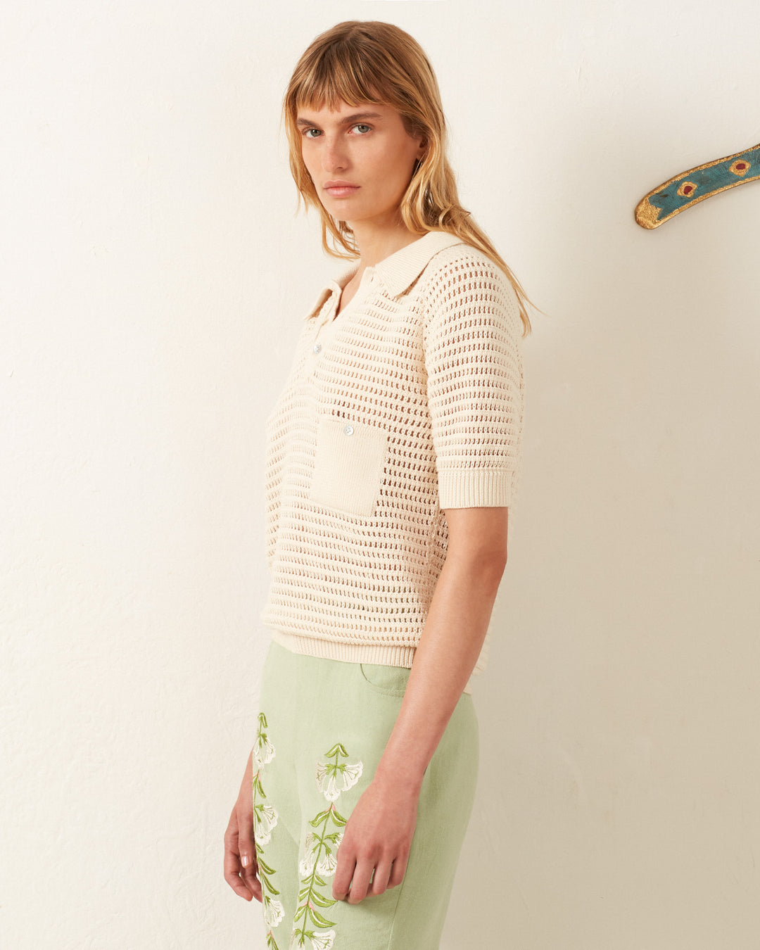 A person in a vintage-inspired Lessi Oyster Crochet Top and a light green skirt, evoking Katharine Hepburn's style, stands against a white wall.