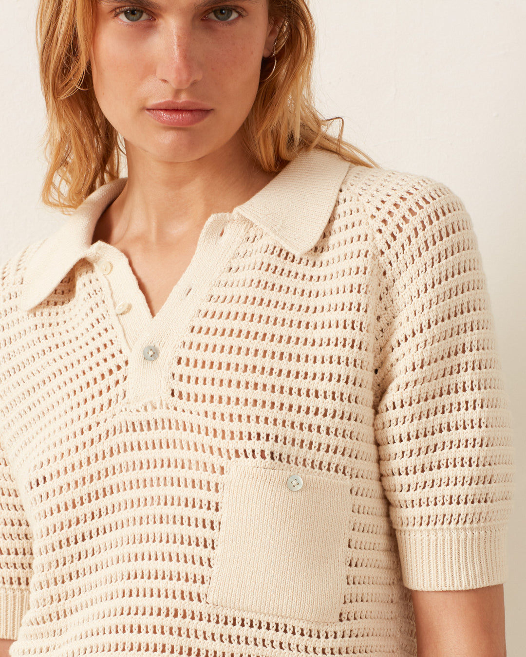 The person is wearing the Lessi Oyster Crochet Top, a vintage-inspired cream knit, in the style of Katharine Hepburn. It features a collar, button placket, short sleeves, and a chest pocket.