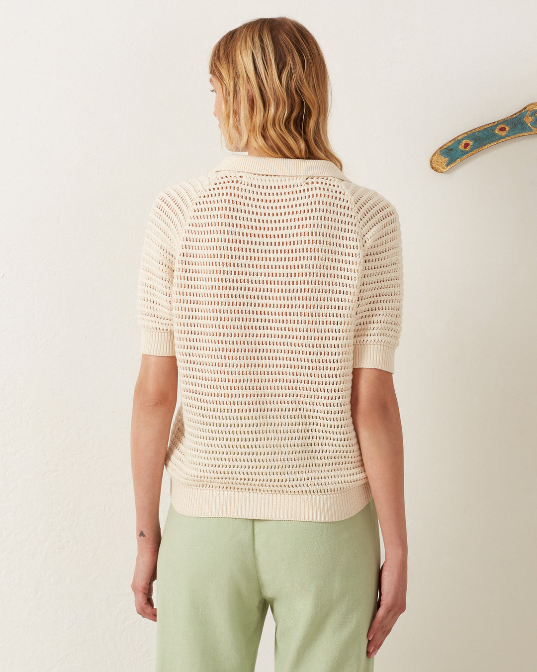 With blonde waves cascading, the person exudes vintage charm in a cream Lessi Oyster Crochet Top and light green pants, subtly channeling Katharine Hepburn's style while they stand with their back to a plain white wall.