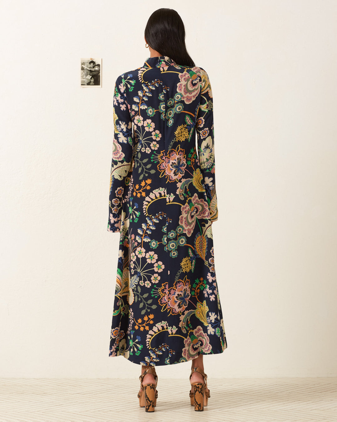 A person in a flowing Nia Desert Flower Dress stands facing a white wall adorned with a small black-and-white photo.